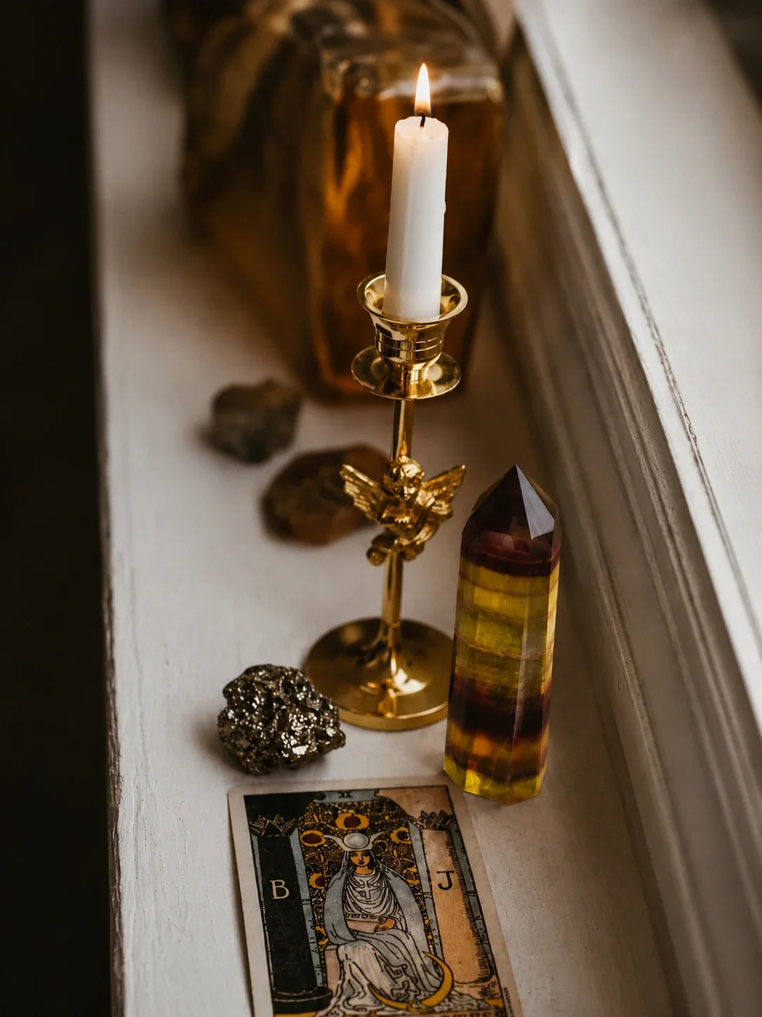 A styled altar-like arrangement on a windowsill featuring a lit white candle in a gold brass holder, a yellow and brown striped crystal tower, a tarot card reading 'The High Priestess,' and small stones against a soft neutral background.