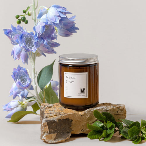 Amber glass jar soy wax candle with silver lid and white label reading Neroli Story, placed on rustic stone with green.