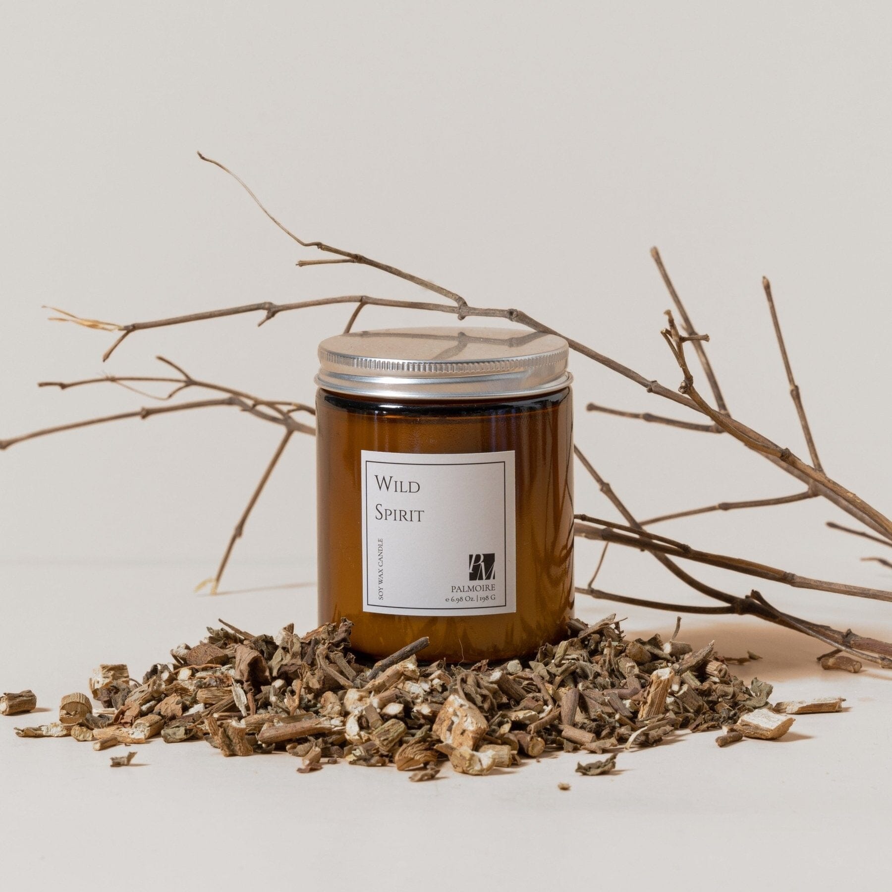 Brown glass jar soy wax candle labeled Wild Spirit surrounded by dried wood chips and bare twigs on white background.