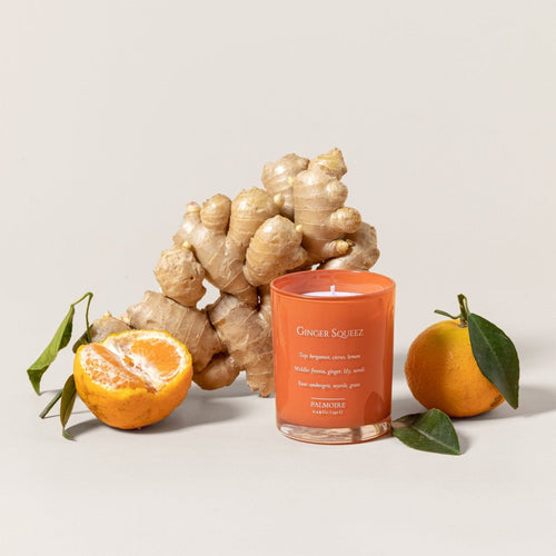 Orange soy wax candle labeled Ginger Squeez with fresh ginger root and whole and halved tangerines with leaves.