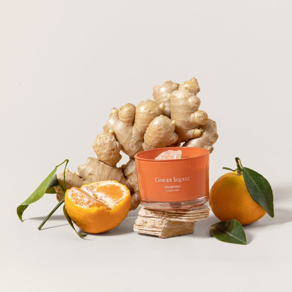 Orange-labeled Ginger Squeez soy wax candle with fresh ginger root, whole and halved mandarin oranges with green leaves.