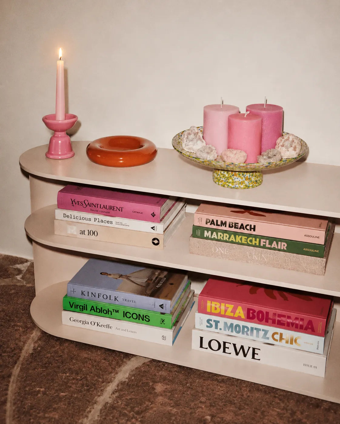 Cream curved bookshelf with books, pink candles, and an orange bowl.