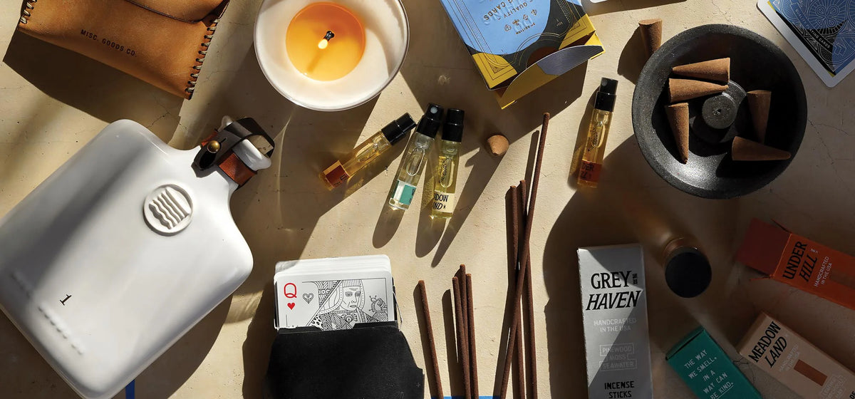 A styled flat-lay of various lifestyle and wellness products on a beige surface, including a white electric hot water bottle, a lit candle, small perfume bottles, chopsticks, a black bowl with wooden sticks, and Grey Haven branded packaging.