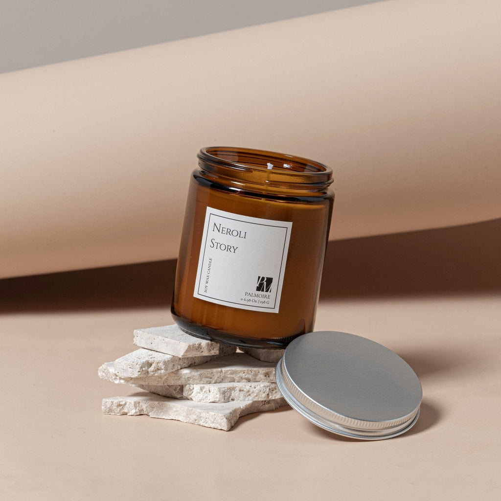 Amber glass jar soy wax candle with white label and silver screw lid on beige stone slabs.