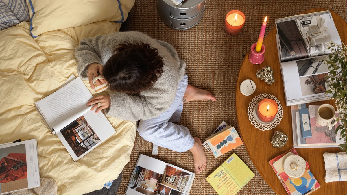 A person sitting on a bed reading a book, surrounded by home decor, candles, and magazines on a woven rug.