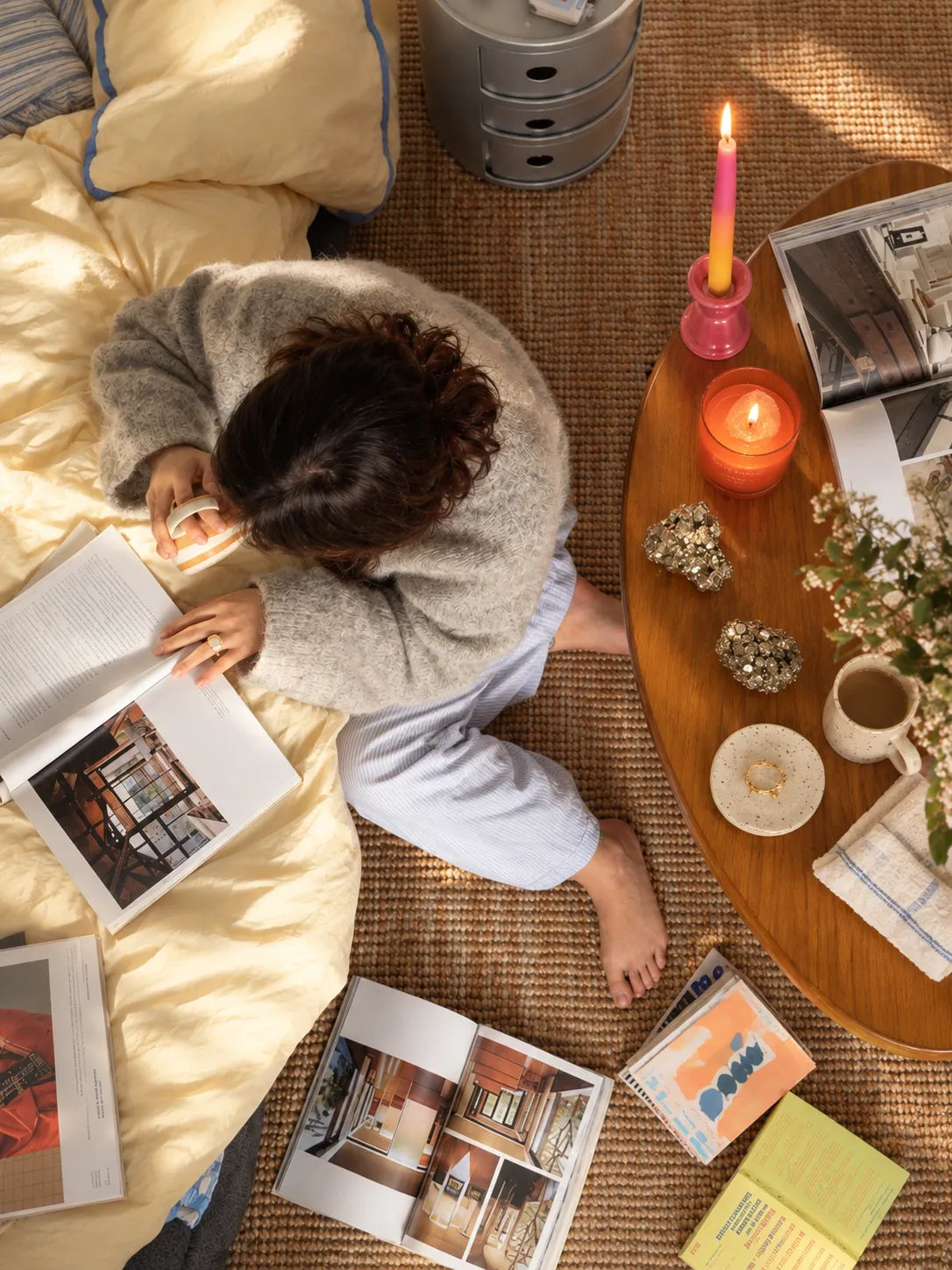 A person sitting on a bed reading a book, surrounded by home decor, candles, and magazines on a woven rug.