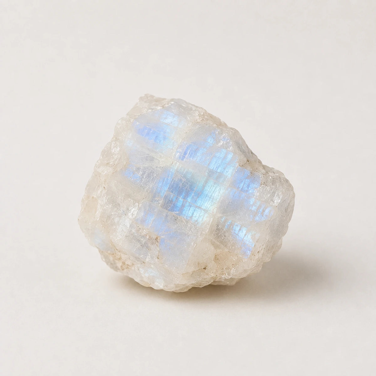 A raw specimen of labradorite crystal showing a soft iridescent blue sheen on a warm off-white base, photographed on a neutral light beige background.