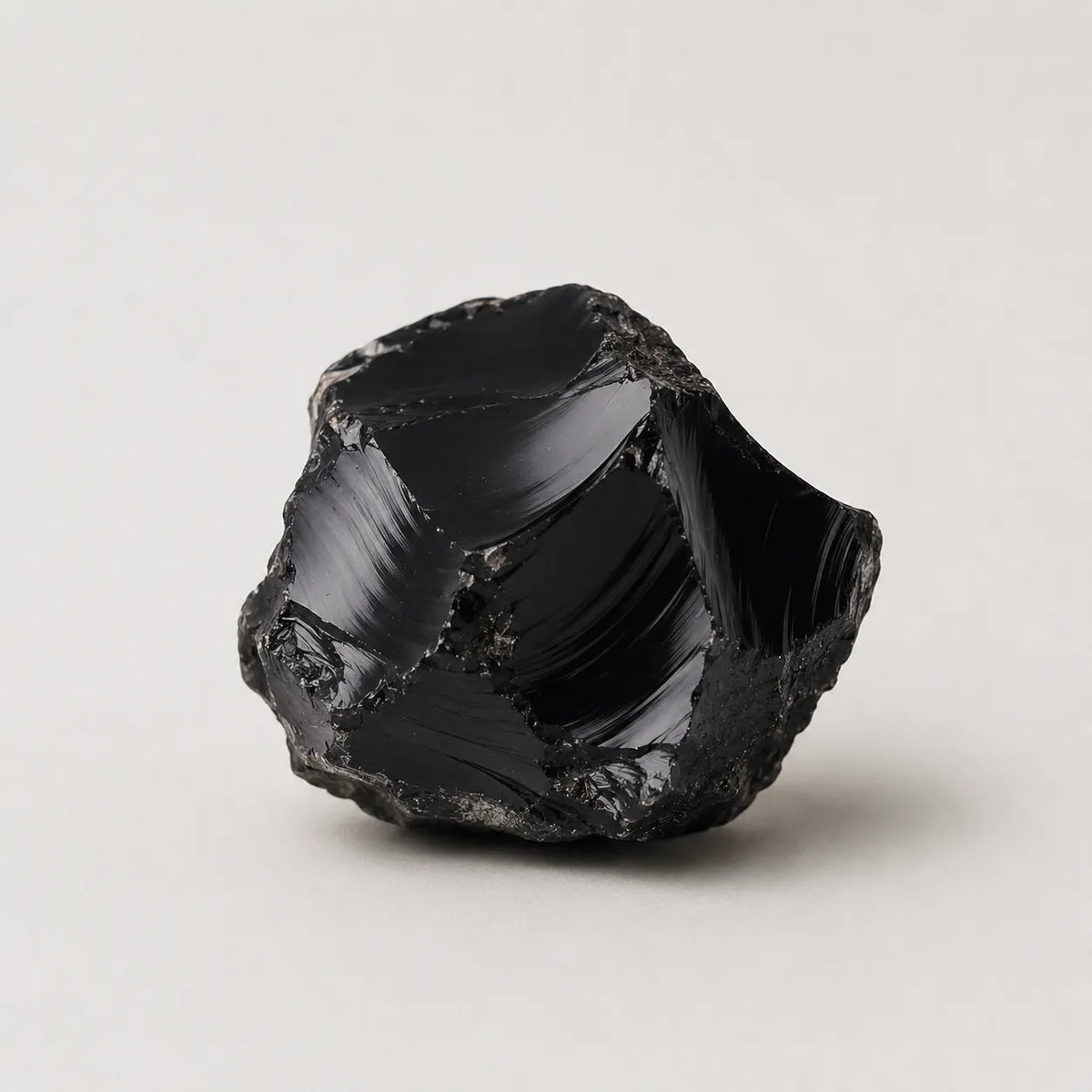 A raw, polished black obsidian stone with sharp, jagged edges and reflective facets, photographed on a neutral light gray background.