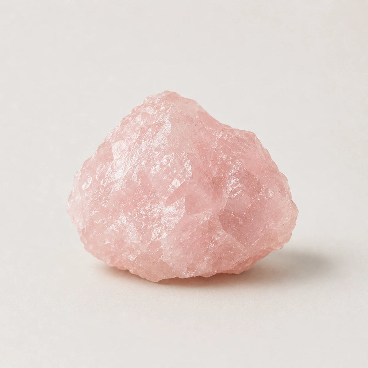 A raw pink rose quartz crystal stone with rough, jagged edges, photographed on a clean, light off-white background.