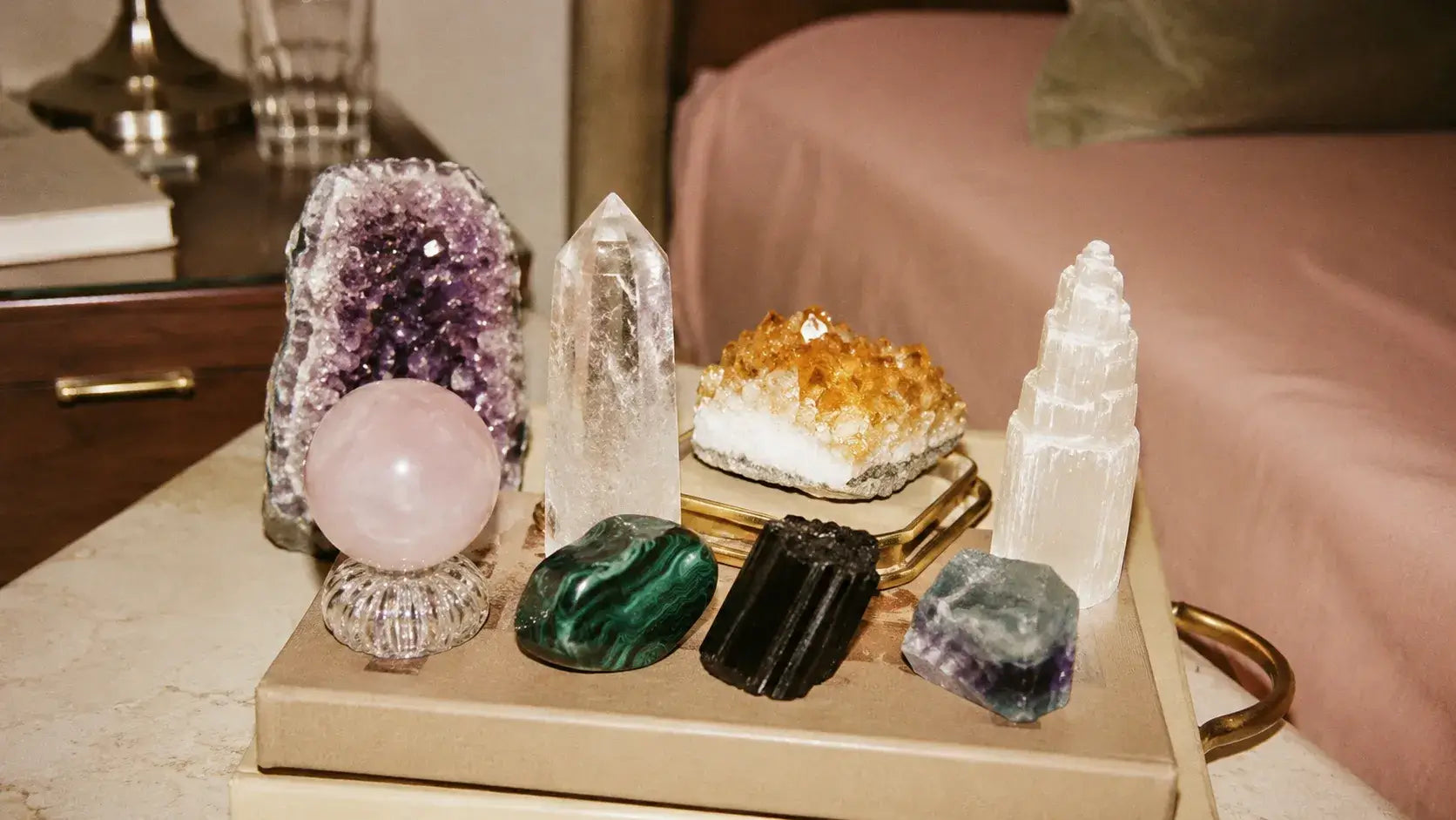 A collection of various crystals and gemstones arranged on a light wood tray on a bedside table, with a bed in the background.