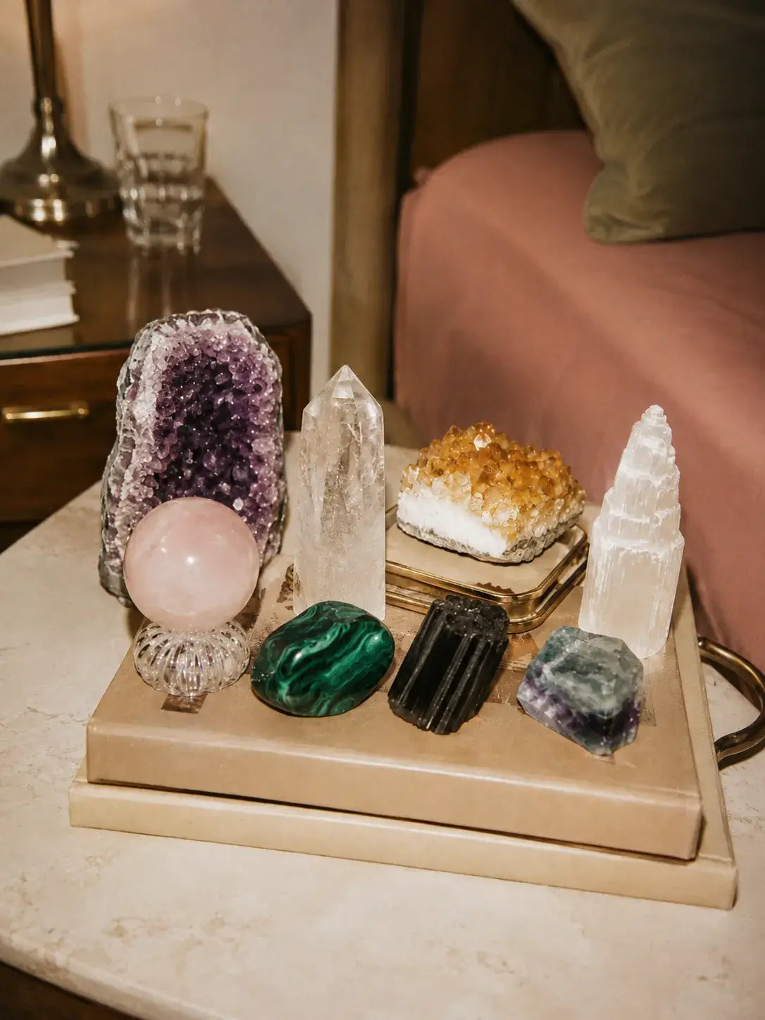 A collection of assorted crystals and gemstones displayed on a beige wooden tray, including a purple amethyst geode, a pink rose quartz sphere, clear quartz points, a green malachite piece, a black bar, and a white selenite tower, set on a bedside table.
