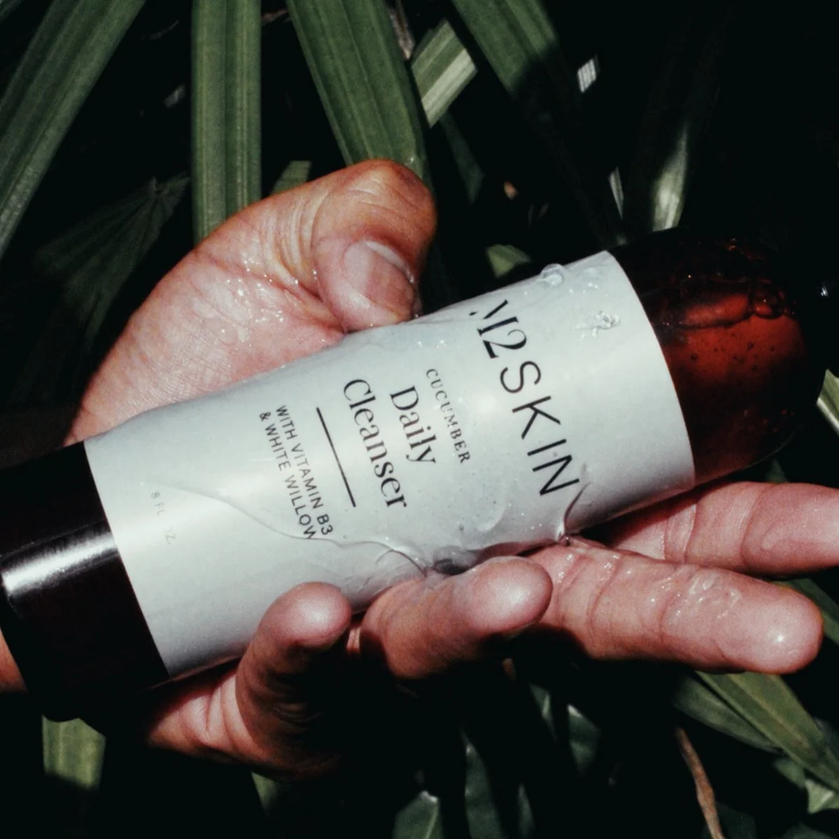 A close-up of a hand holding a bottle of M2 SKIN Cucumber Daily Cleanser with Vitamin B3 and White Willow against a tropical leaf background.