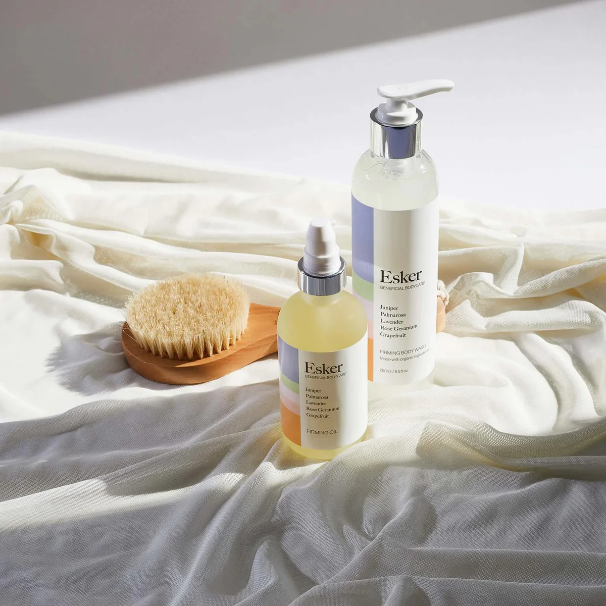 Esker firming body oil and wash presented with a wooden dry brush on a white fabric for a luxury body care routine.