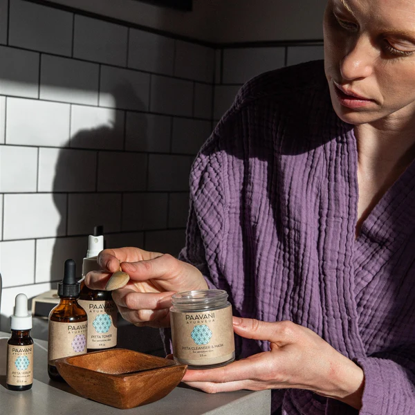 A woman scoops herbal powder from a Paavani Ayurveda Pitta Cleanser & Mask jar to prepare a natural face mask for sensitive skin.