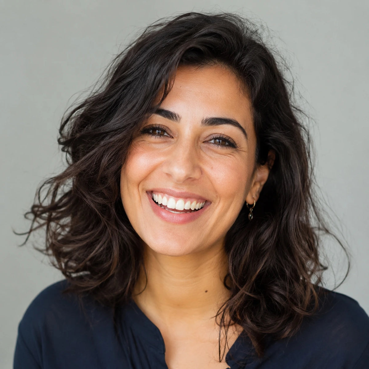 A smiling woman with dark wavy hair wearing a navy top, photographed in a close-up portrait against a plain light gray background.