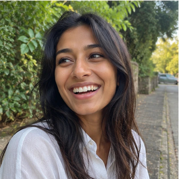 A smiling woman with long dark hair wearing a white button-up shirt, photographed outdoors on a paved path with green foliage and trees in the background.