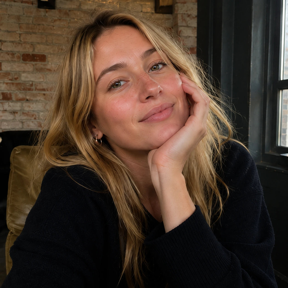 A close-up portrait of a woman with blonde hair resting her head on her hand, wearing a dark navy knit sweater, set against an indoor background with a brick wall and window.