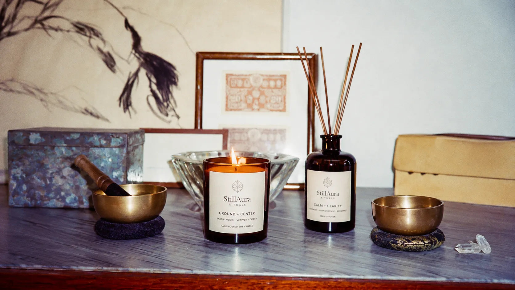 A styled product display featuring a lit candle and a reed diffuser bottle from the brand StillAura, set on a wooden table alongside brass singing bowls, a glass bowl, and framed art in a warm, atmospheric setting.